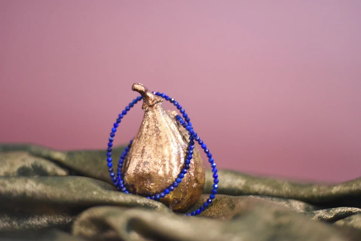 Ordoñez Le blue beaded necklace draped over a gold-colored pear on green fabric against a pink backdrop, from the official brand website